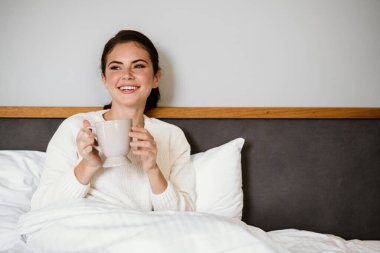 Sevimli neşeli kız evde kötü otururken gülümsüyor ve kahve içiyor.