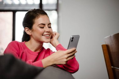 Cep telefonu tutan mutlu genç bir kadın evdeki kanepede oturuyor.