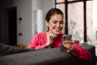 Cep telefonu tutan mutlu genç bir kadın evdeki kanepede oturuyor.