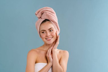 Havlu giymiş neşeli ve çekici bir kız mavi arka planda izole edilmiş kamera önünde poz veriyor ve gülümsüyor.