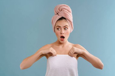 Havlu giymiş beyaz bir kız mavi arka planda izole edilmiş kameraya şaşkınlığını dile getiriyor.
