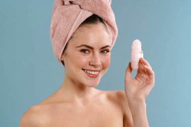 Mavi arka planda izole edilmiş kozmetik süngerle poz veren havluyla gezen gömleksiz neşeli kız.