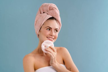 Mavi arka planda izole edilmiş kozmetik süngerle poz veren mutlu bir kız.