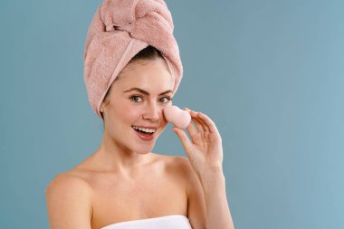 Mavi duvar arkasında izole edilmiş havluyla makyaj süngeri tutan mutlu genç bir kadın resmi