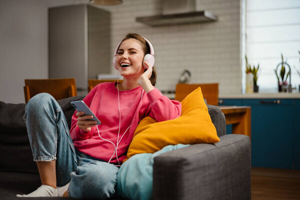 Nice joyful girl listening music with headphones and cellphone while sitting on couch at home