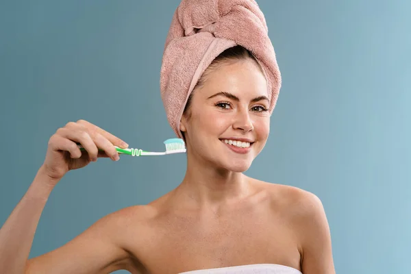 漂亮而快乐的女孩穿着毛巾 拿着牙刷 牙膏与蓝色背景隔离 — 图库照片