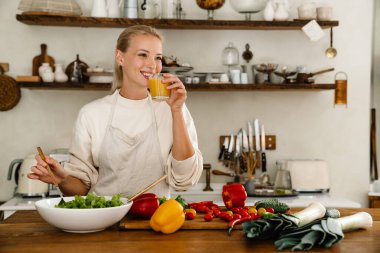 Güzel, mutlu bir kadın. Mutfakta yemek yaparken gülümsüyor ve meyve suyu içiyor.