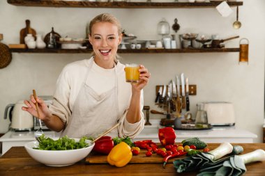 Güzel, mutlu bir kadın. Mutfakta yemek yaparken gülümsüyor ve meyve suyu içiyor.