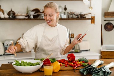Güzel, mutlu bir kadın. Mutfakta yemek yaparken cep telefonu kullanıyor ve gülümsüyor.