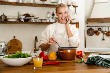 Güzel, mutlu bir kadın. Mutfakta yemek hazırlarken cep telefonuyla konuşuyor.