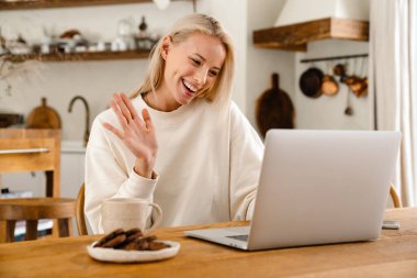Güzel, neşeli bir kadın çay içip el sallarken rahat bir mutfakta dizüstü bilgisayarla çalışıyor.