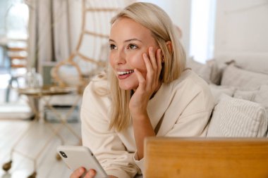 Güzel, mutlu bir kadın rahat bir odada kanepede uzanırken gülümsüyor ve cep telefonu kullanıyor.