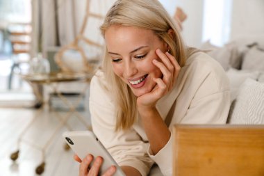 Güzel, mutlu bir kadın rahat bir odada kanepede uzanırken gülümsüyor ve cep telefonu kullanıyor.