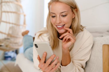 Güzel, neşeli bir kadın rahat bir odada kanepede uzanırken gülümsüyor ve cep telefonu kullanıyor.