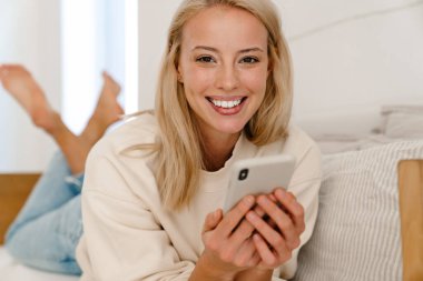 Güzel, mutlu bir kadın rahat bir odada kanepede uzanırken gülümsüyor ve cep telefonu kullanıyor.