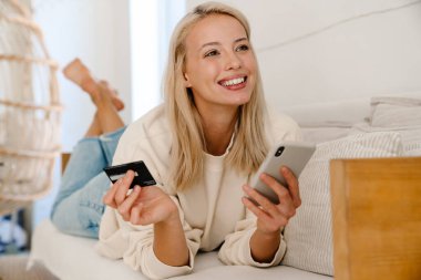 Güzel, neşeli kadın rahat bir odada kanepede uzanırken kredi kartı ve cep telefonu kullanıyor.