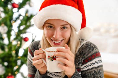 Noel Baba şapkalı güzel bir kadın elinde arka planda Noel ağacı olan sıcak bir içecek tutuyor.