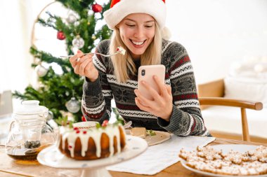 Noel Baba şapkalı mutlu bir genç kızın evde cep telefonu kullanırken pasta yemesi.