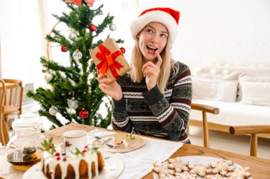 Noel Baba şapkası takan düşünceli güzel bir kadın Noel 'i çam ağacıyla kutluyor ve elinde hediye kutusuyla pasta tutuyor.
