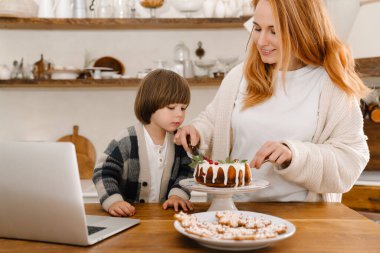 Mutlu anne ve oğul Noel pastası yerken ev mutfağında dizüstü bilgisayar kullanıyor.