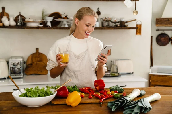 Beautiful happy woman using cellphone and drinking juice while making lunch in cozy kitchen