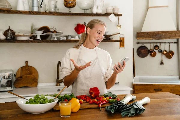 Güzel, mutlu bir kadın, rahat bir mutfakta yemek yaparken cep telefonu ve dolma biber kullanıyor.