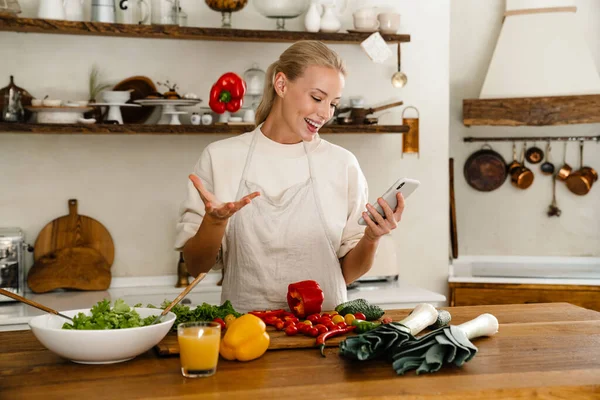 Güzel, mutlu bir kadın, rahat bir mutfakta yemek yaparken cep telefonu ve dolma biber kullanıyor.