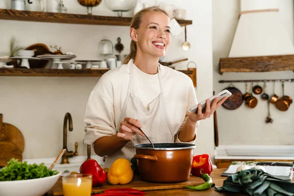 Mulher Alegre Bonita Sorrindo Usando Celular Enquanto Faz Almoço Cozinha — Fotografia de Stock