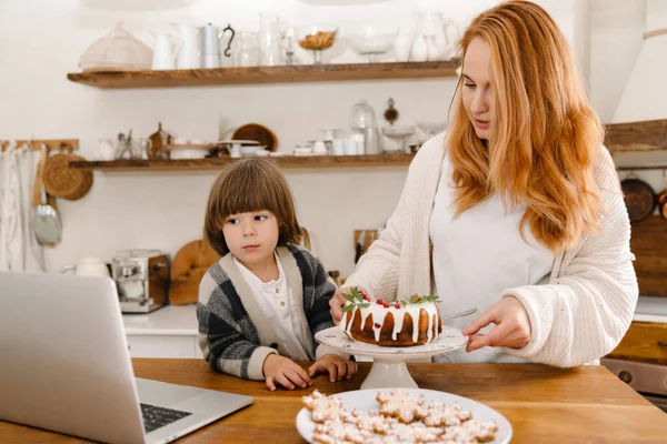 一个乐观的年轻母亲和她的儿子一起在家里用笔记本电脑做饭和做圣诞蛋糕的照片 — 图库照片