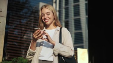 Çekici, genç, sarışın bir kadın binanın yanındaki sokakta akıllı telefonunu kullanıyor.