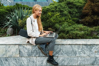 Dışarıda dizüstü bilgisayar kullanarak oturan mutlu ve çekici genç kadın.