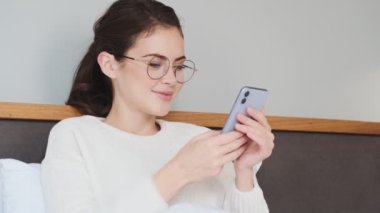 Genç güzel kız yatakta uzanıyor ve cep telefonunu evde kullanıyor.