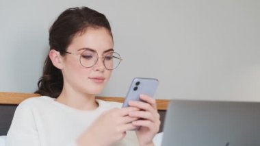 Genç güzel kız yatakta uzanıyor ve cep telefonunu evde kullanıyor.