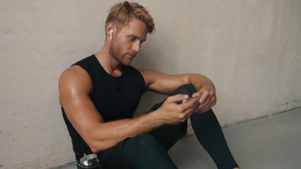 一个拿着耳机的快乐男人正在用他的智能手机横向地拿着耳机 同时坐在里面灰色的墙上 — 图库视频影像