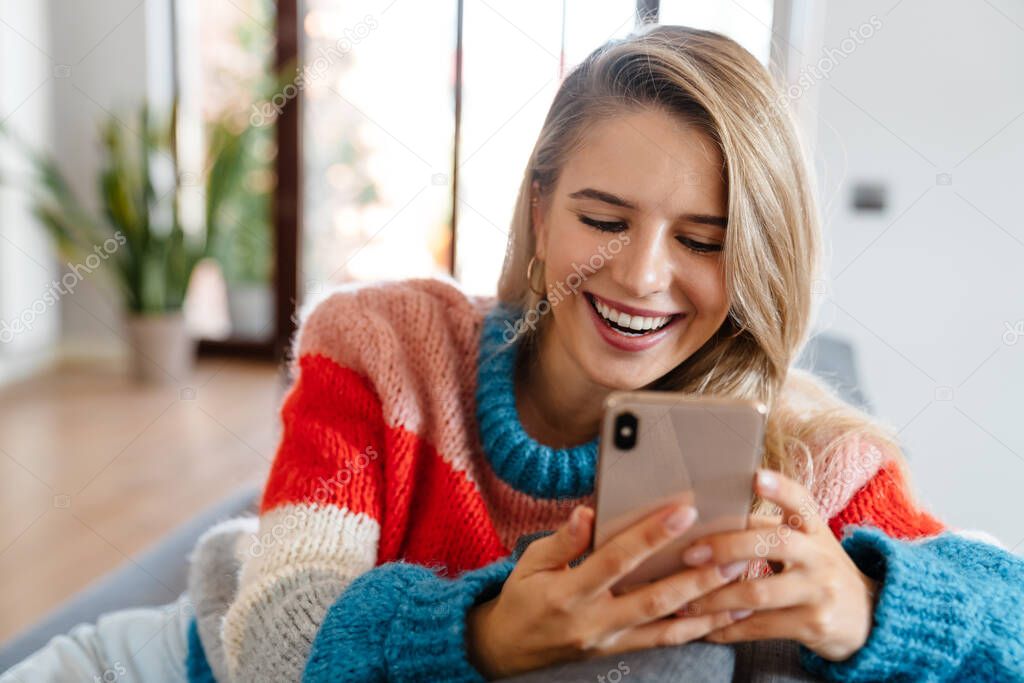 Attractive smiling young woman using mobile phone while ralaxing on couch at home