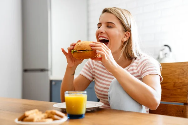 笑顔若いです女性食べるハンバーガーにキッチンに自宅 — ストック写真