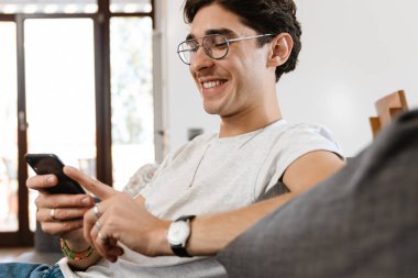 Evdeki koltukta oturup cep telefonu kullanan mutlu iyimser bir adamın fotoğrafı.