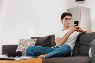 Evdeki kanepede oturan yakışıklı adamın fotoğrafı. Cep telefonu kullanırken.