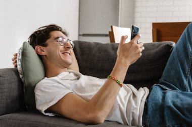 İyimser memnun yakışıklı adamın resmi evdeki kanepede cep telefonuyla yatıyor.