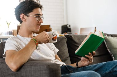Evde çay içip kitap okuyan yakışıklı bir adamın resmi.