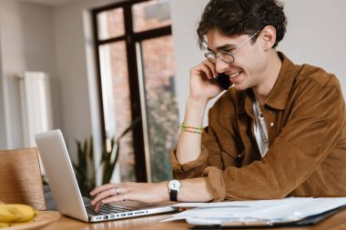 Evde cep telefonuyla konuşan ve dizüstü bilgisayar kullanan neşeli bir adamın fotoğrafı.
