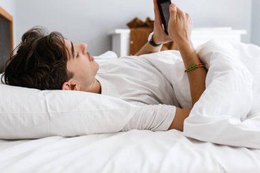 Yakışıklı bir adam yatakta uzanıyor ve cep telefonunu evde kullanıyor.