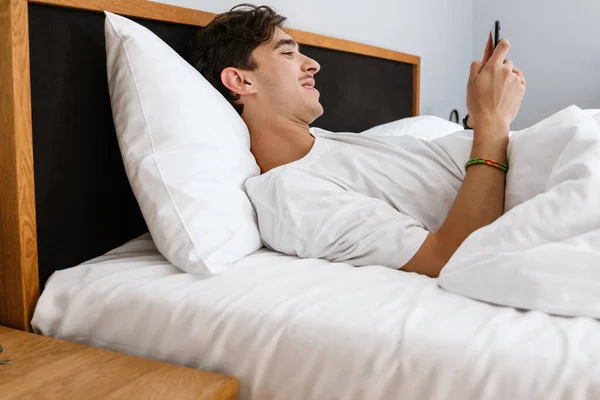 Imagen Del Hombre Complacido Encuentra Cama Uso Teléfono Móvil Interior —  Fotos de Stock