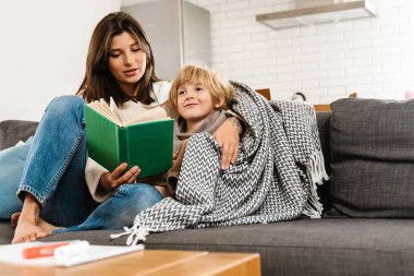 Evinde oğlu için kitap okuyan pozitif gülümseyen genç bir kadın resmi.