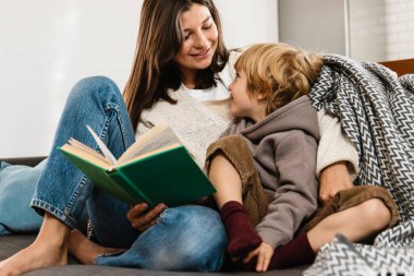 Evinde oğlu için kitap okuyan güzel bir kadın resmi.