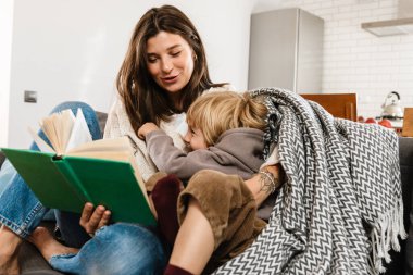 Evinde oğlu için kitap okuyan güzel bir kadın resmi.