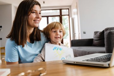 Genç ve güzel bir kadının, oğluyla evde çekilmiş fotoğrafı. Çizimlerini dizüstü bilgisayarla gösteriyor.