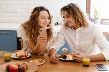 Mutlu, güzel bir çift, gülümsüyor ve rahat bir mutfakta kahvaltı ederken cep telefonu kullanıyor.