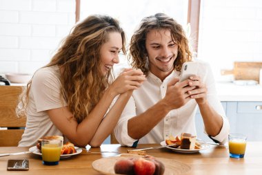 Mutlu, güzel bir çift, gülümsüyor ve rahat bir mutfakta kahvaltı ederken cep telefonu kullanıyor.