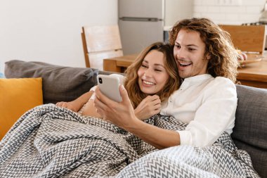 Mutlu, güzel bir çift gülümsüyor ve evdeki kanepede dinlenirken cep telefonu kullanıyor.